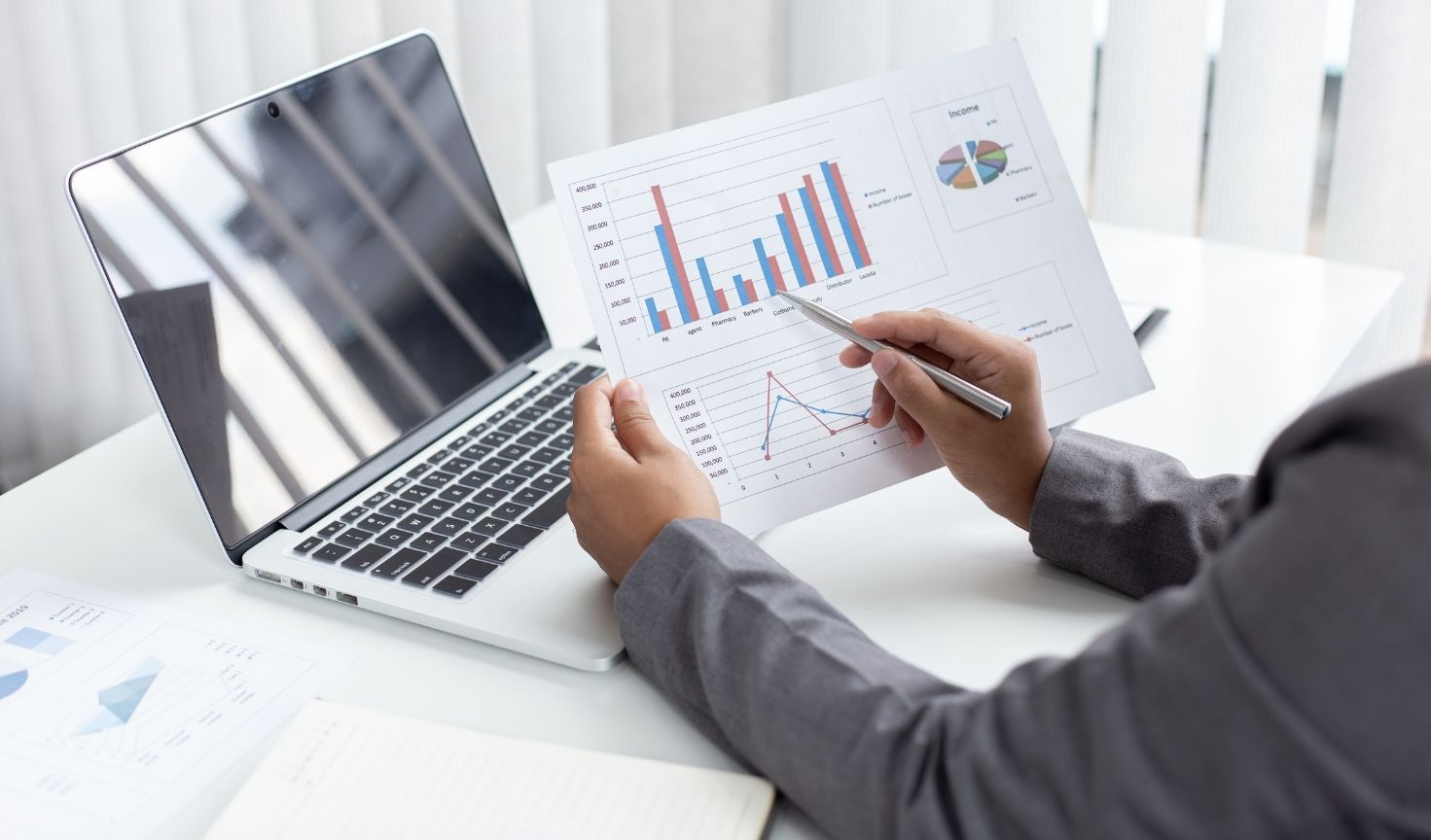 Person in business attire reviewing printed financial charts and graphs while sitting at a desk with an open laptop, pointing at the data with a pen.