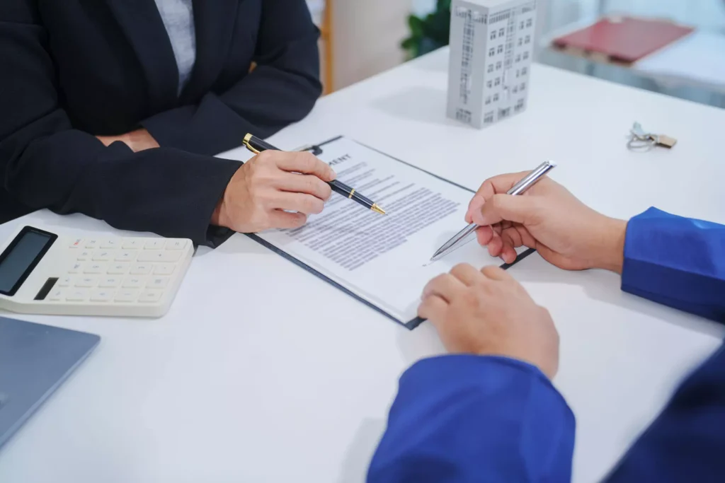 Two people in business attire reviewing and pointing to a printed contract on a desk, with pens in hand and a calculator nearby, indicating a financial or property agreement discussion.