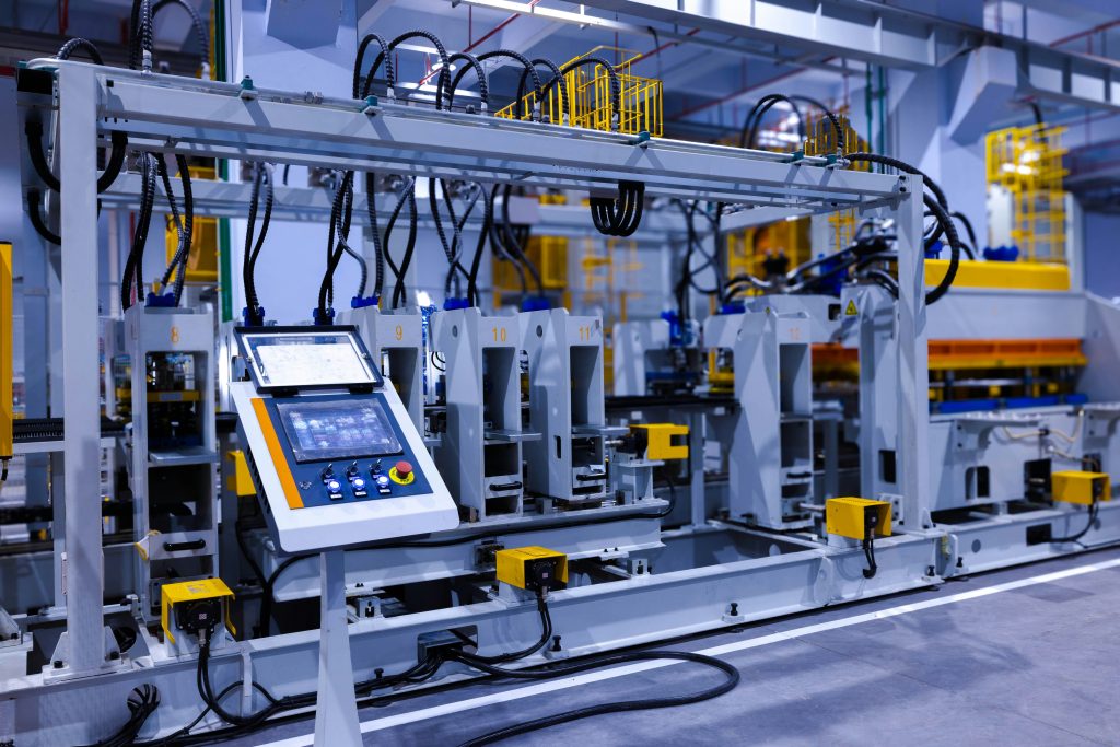 Automated industrial production line with robotic stations, control panel screen, and overhead cabling inside a modern factory facility.