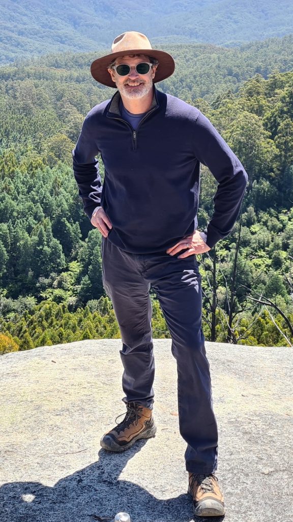 Man wearing a wide-brim hat and sunglasses standing on a rocky lookout with hands on hips, overlooking a vast forested mountain landscape under a clear blue sky.