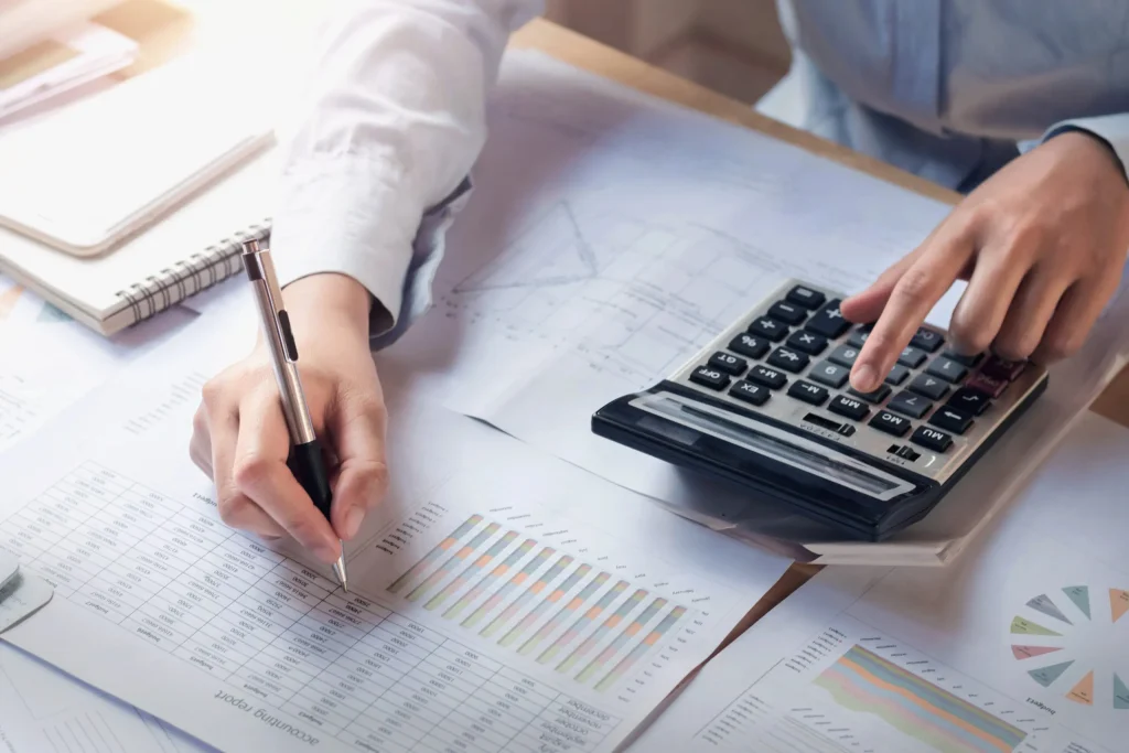 Close-up of hands reviewing financial documents, one hand pointing to a spreadsheet with a pen while the other presses buttons on a calculator.