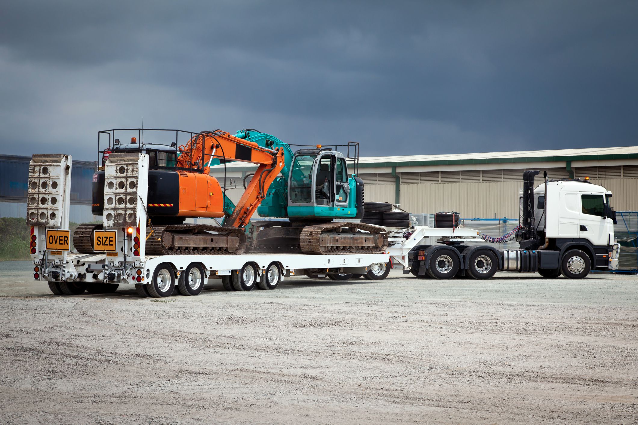 Flatbed truck transporting two tracked excavators at an industrial yard under a cloudy sky, with “oversize” warning signs on the trailer.