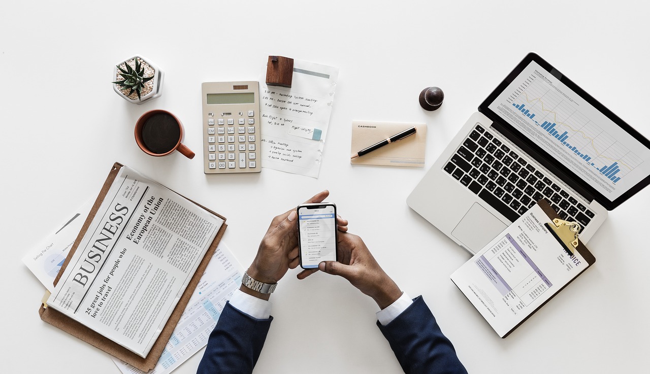 Top-down view of a tidy office desk with a person holding a smartphone, surrounded by a laptop displaying financial charts, calculator, notebook, coffee cup, newspaper, and business documents.