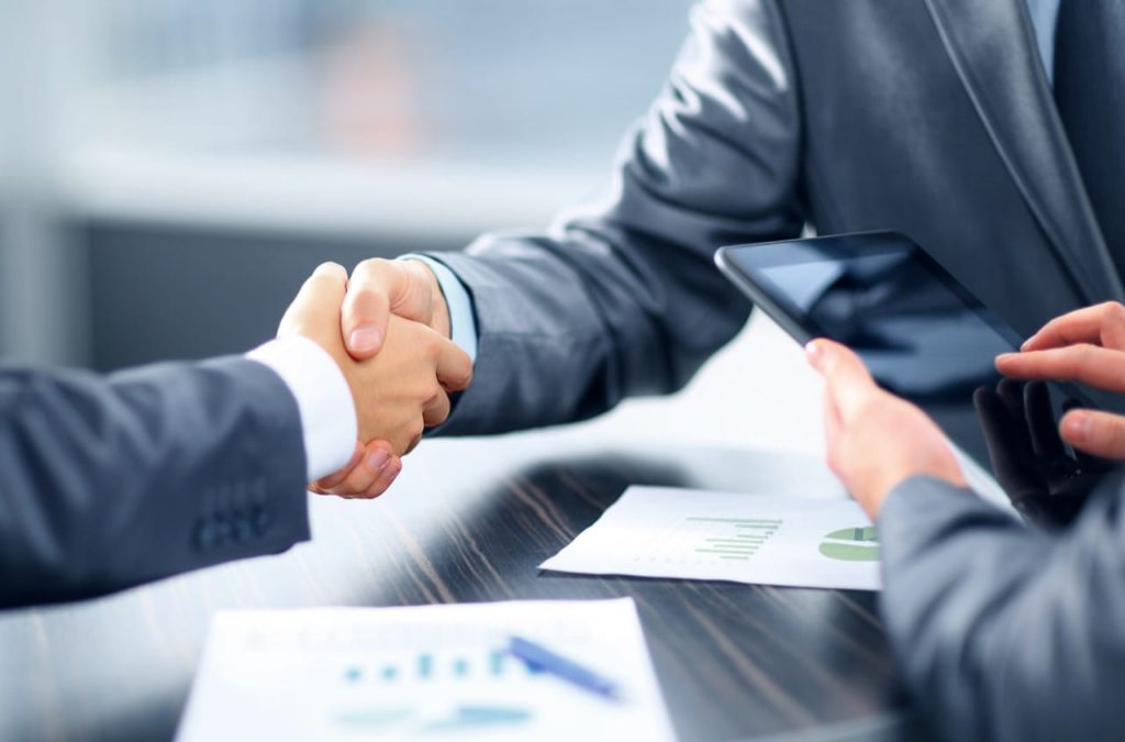 Two people in business suits shaking hands over a desk with a small model house in the foreground, while another person holds out a set of keys, suggesting a property sale or real estate agreement.