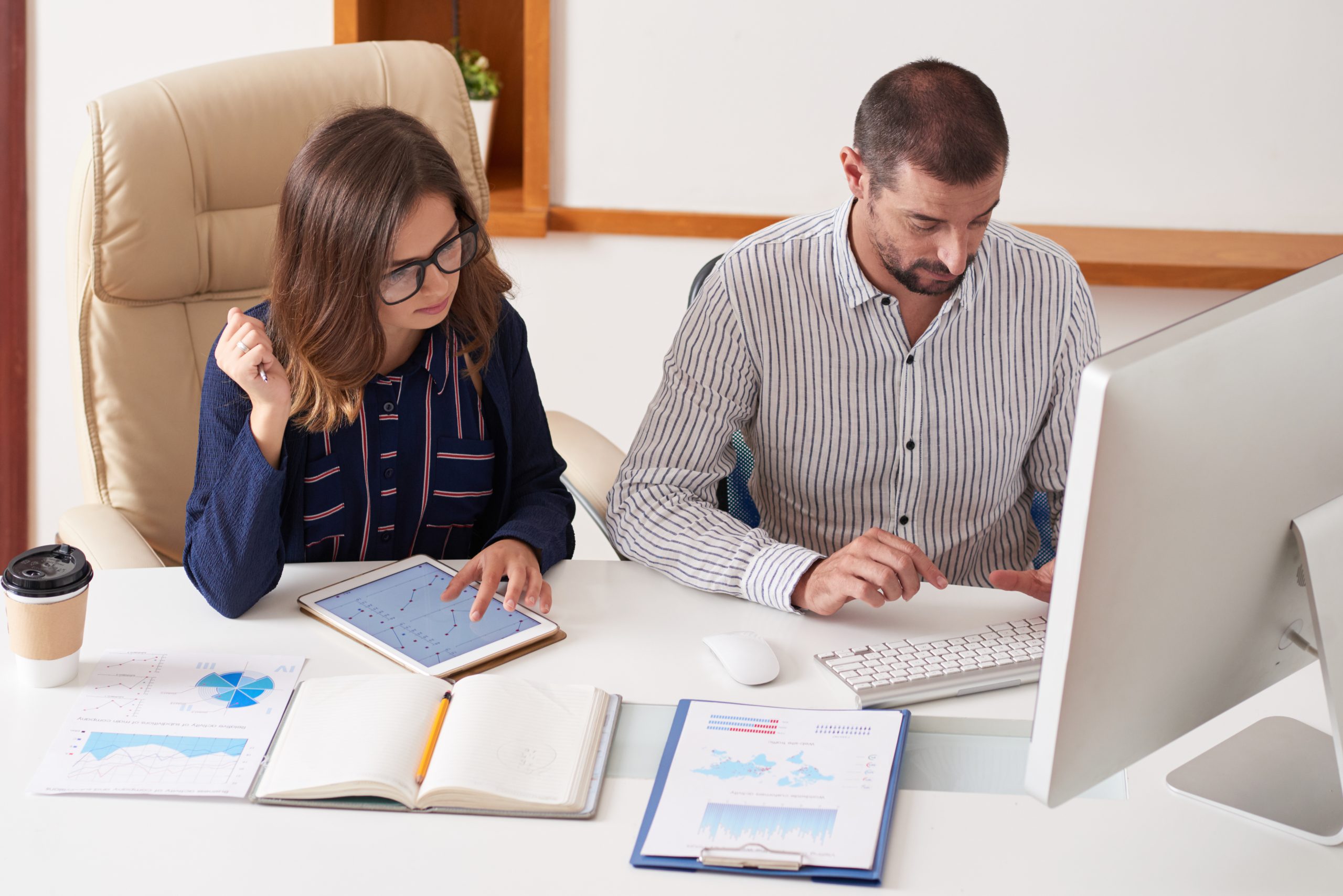 Two colleagues sitting at a desk analysing financial data, one using a tablet with charts while the other works on a desktop computer, with reports and a notebook spread out on the table.
