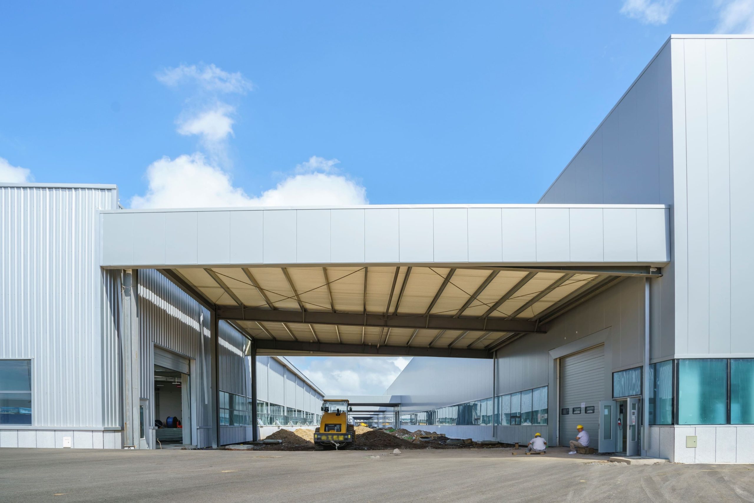 Industrial warehouse buildings connected by a covered loading area, with a small construction vehicle and workers near piles of dirt in an open concrete yard