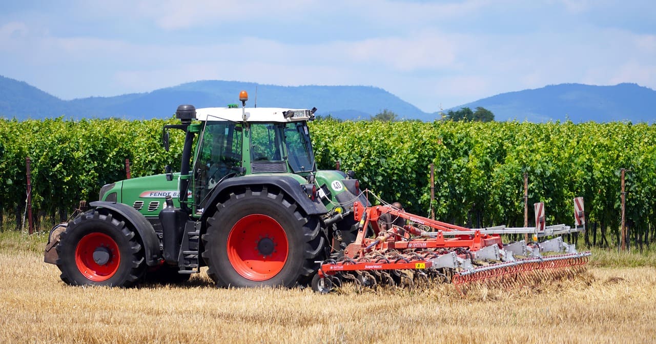Green tractor pulling a large tillage implement across a dry field beside rows of vineyard plants, with rolling hills and mountains in the background
